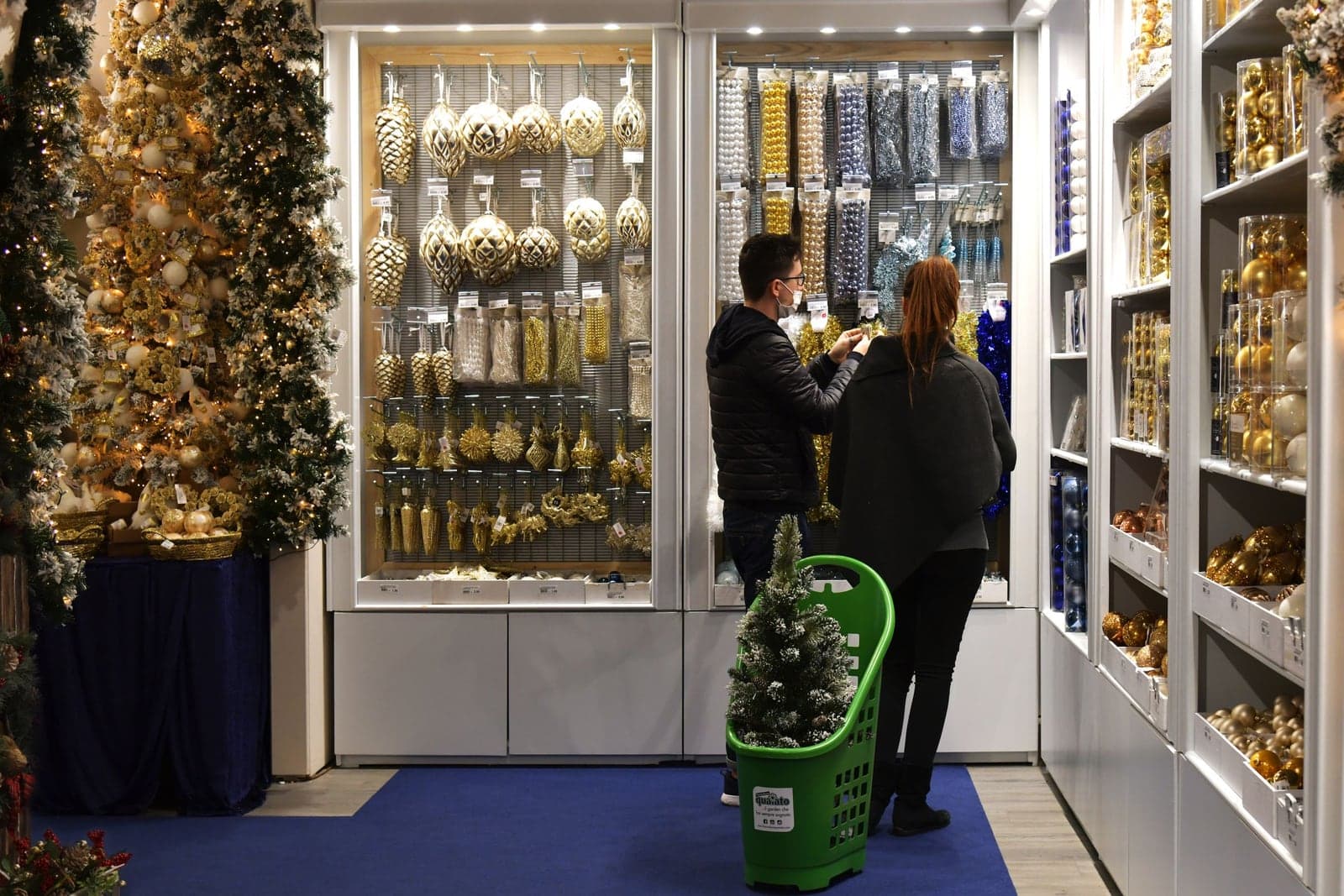 Two shoppers browsing a wall of gold and silver Christmas ornaments and garland in a brightly lit holiday decoration store, with flocked trees and bins of baubles lining the aisles