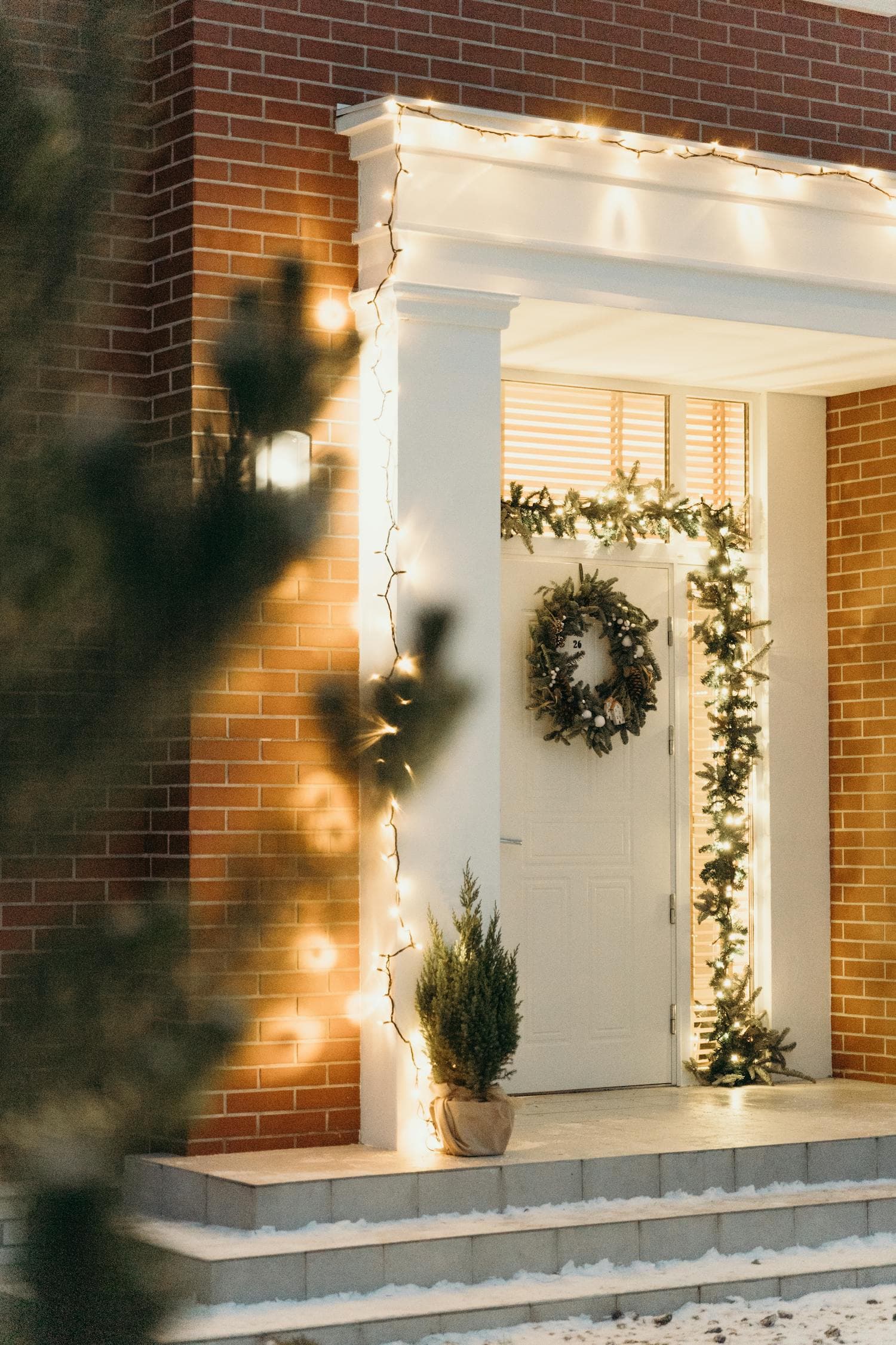 A white-columned brick-home entry with warm-white string lights wrapped from base to capital along the porch column, evergreen garland framing the front door, and a lit wreath on the door — the column-wrapping move that defines a Craftsman porch at Christmas
