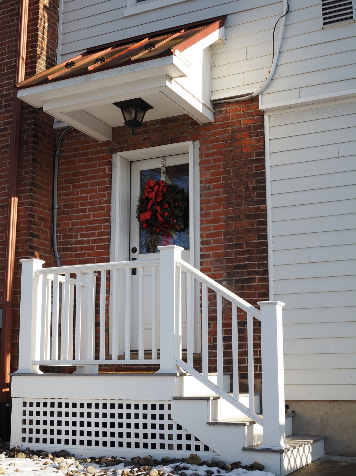 A small white-railed covered porch on a brick-and-clapboard home with a single restrained wreath and red bow on the front door — the restraint-first approach that defines modern-farmhouse porch decor