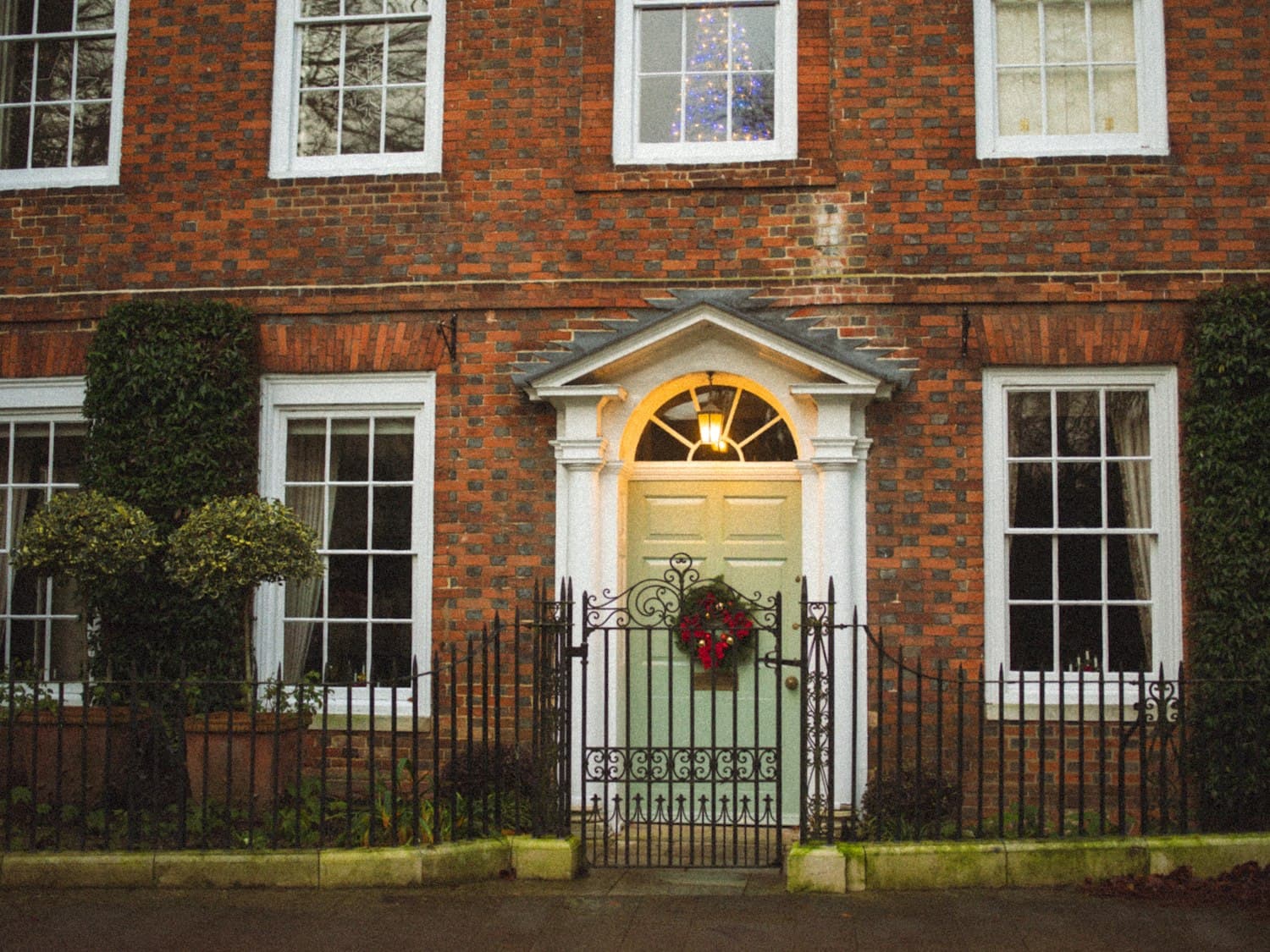 A Georgian brick home with a sage-green front door, classical pediment overhead, and a single Christmas wreath with red ornaments sized to roughly 60% of the door's width — a textbook example of decor that frames the door instead of burying it, plus a Christmas tree visible through the upstairs window