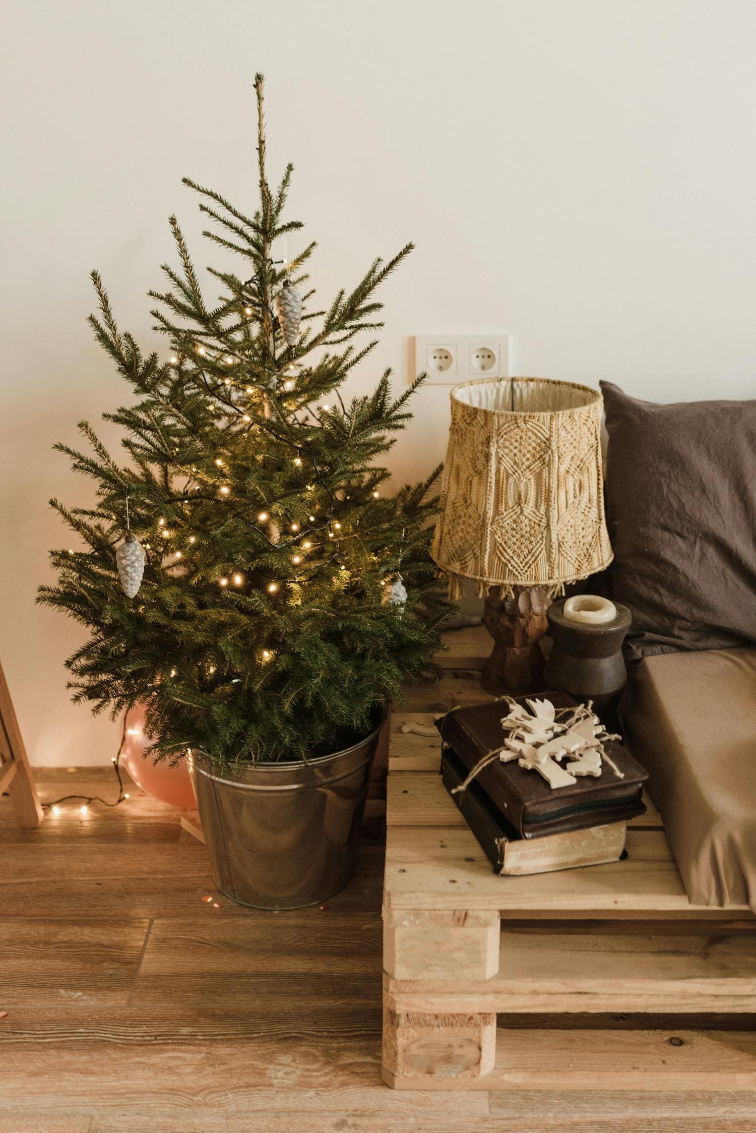 Small live Christmas tree in a galvanized bucket wrapped in warm-white string lights, beside a taupe sofa and a wooden pallet side table with a macrame lamp — a cozy small-apartment Christmas corner