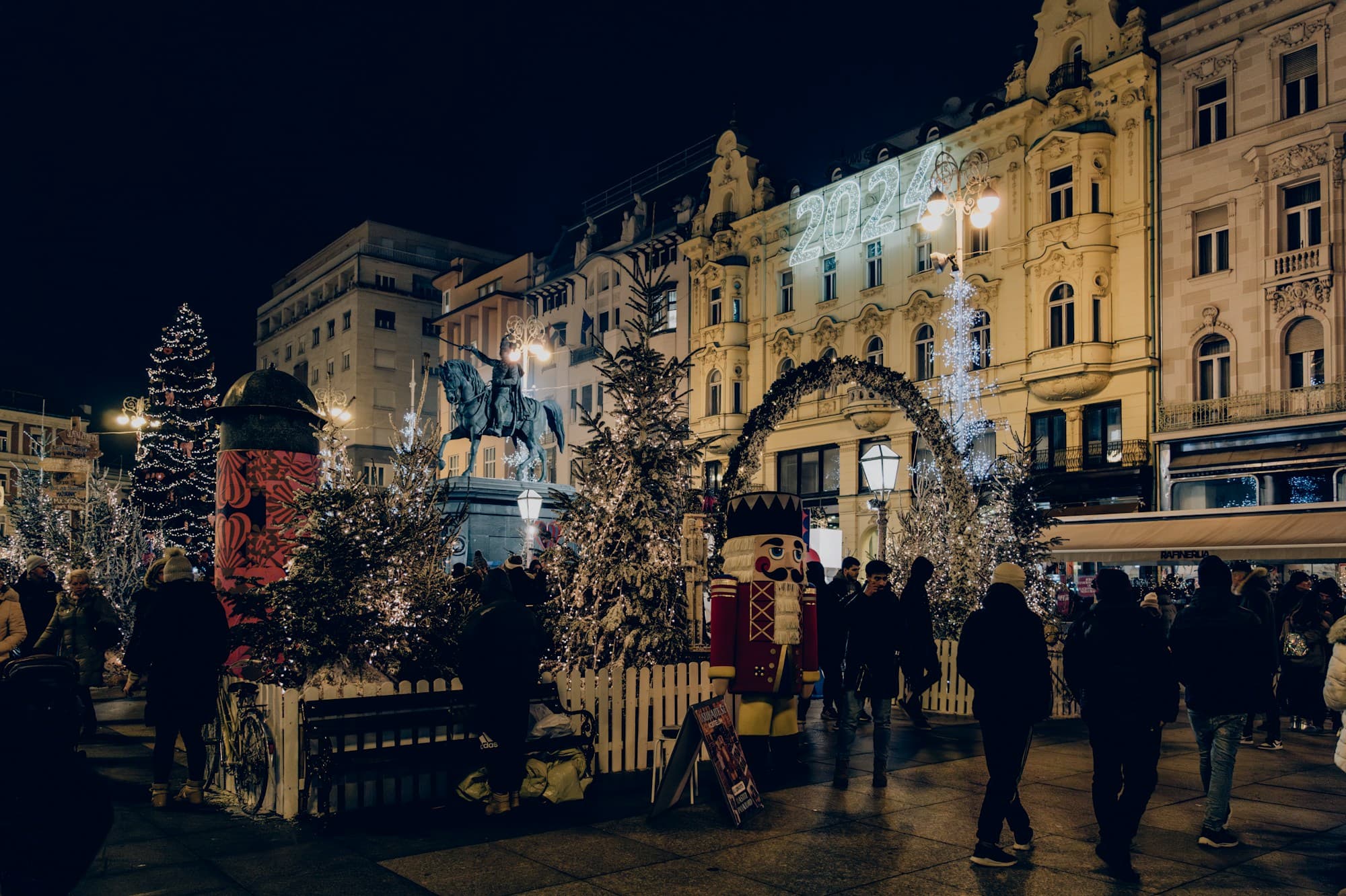 Zagreb's main square during Advent at night, with illuminated Christmas trees, a giant nutcracker, a lit archway, a "2024" light display on a grand belle-epoque building, the equestrian statue of Ban Jelačić, and crowds of visitors among the festive lights — Zagreb's repeatedly award-winning Christmas market