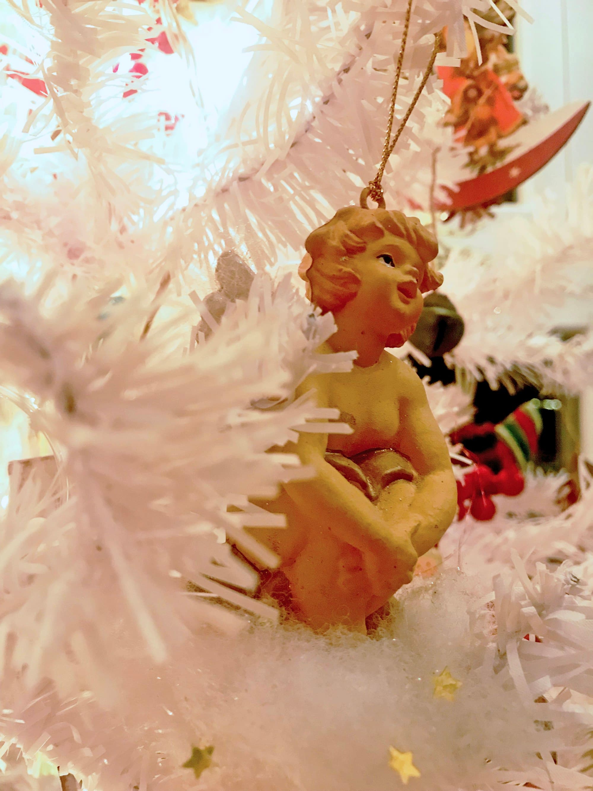 A small cherub ornament with golden curls hangs by a gold thread among the white frosted branches of a Greek Christmas tree, photographed close in Athens with warm interior light filtering through the branches