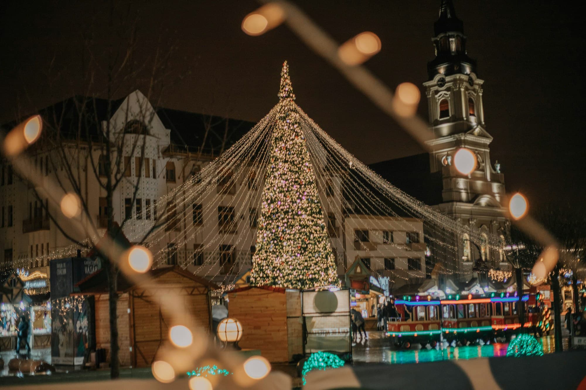 A large illuminated Christmas tree in a Romanian town square at night, with light streamers cascading from its peak down to the surrounding wooden Christmas market stalls, an Orthodox church bell tower visible to the right, framed through string-light bokeh in the foreground