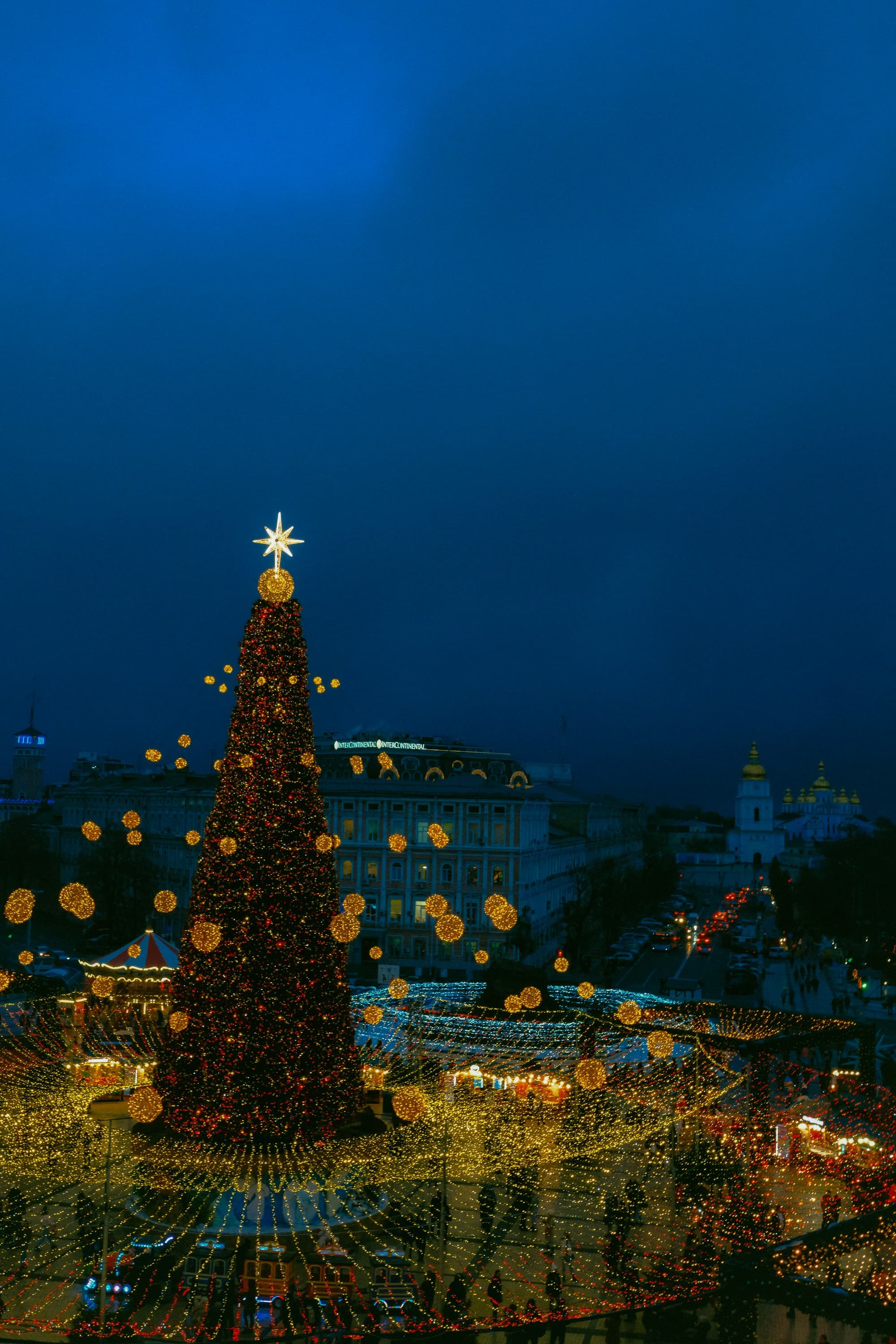 An enormous illuminated Christmas tree topped with a golden Star of Bethlehem in Kyiv's Sofiyivska Square, surrounded by golden bauble decorations strung in concentric rings, with St. Michael's Golden-Domed Cathedral visible in the background against a deep blue twilight sky