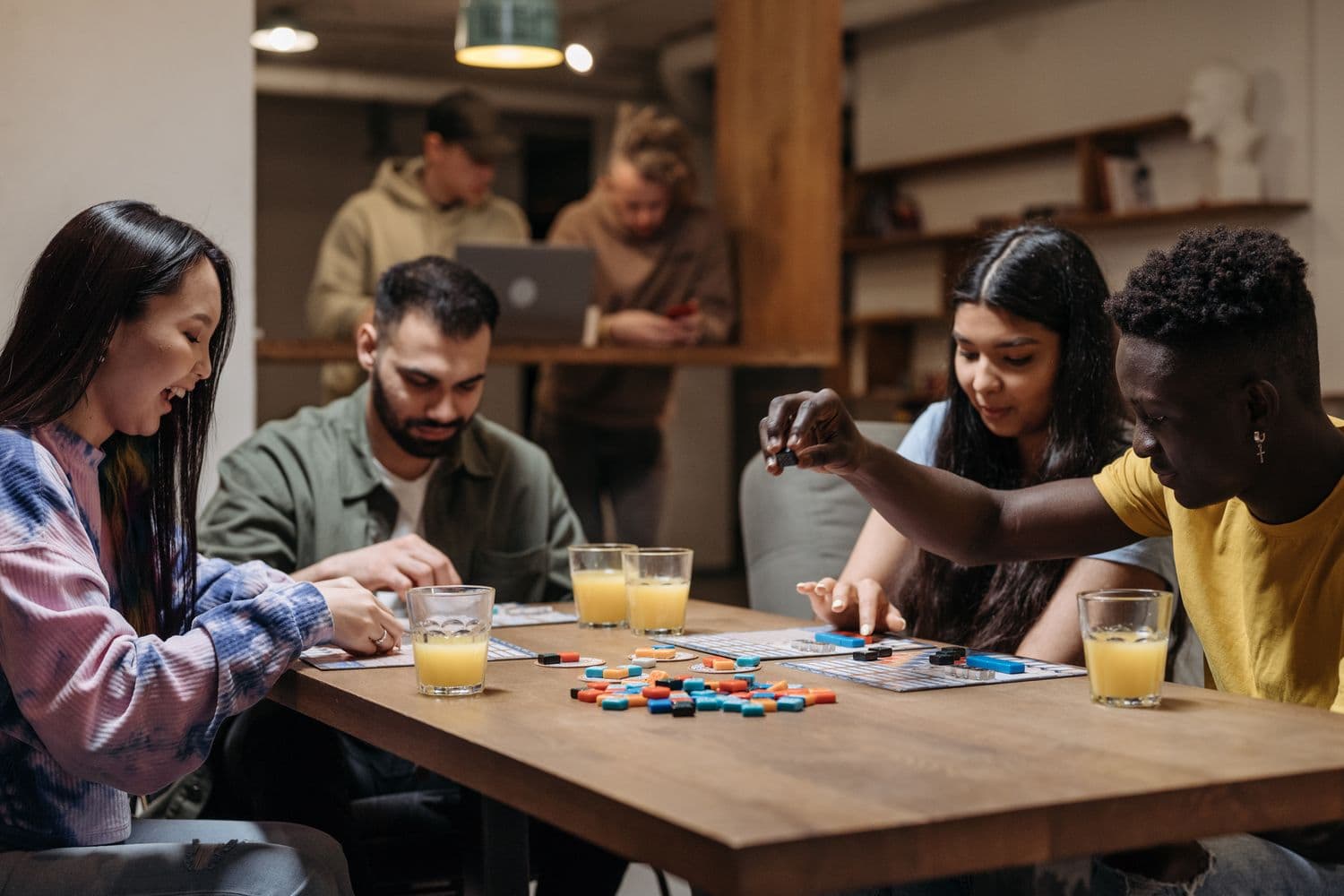 A group of friends gathered around a table playing a tabletop game together with drinks, laughing in a warm, low-lit room