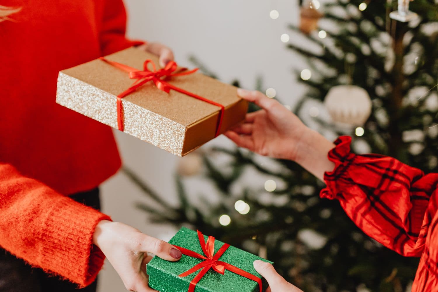 Two people exchanging small wrapped gifts in front of a lit Christmas tree, one gold and one green present with red ribbons