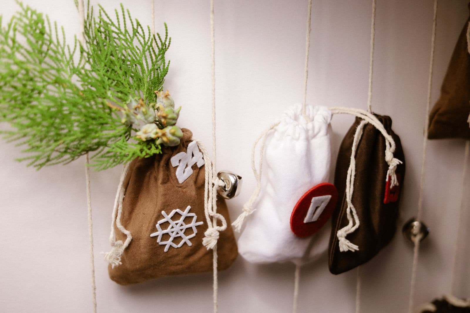 A row of small fabric drawstring pouches in brown, cream, and dark green hanging from twine against a pale wall, with a sprig of cedar and a small bell