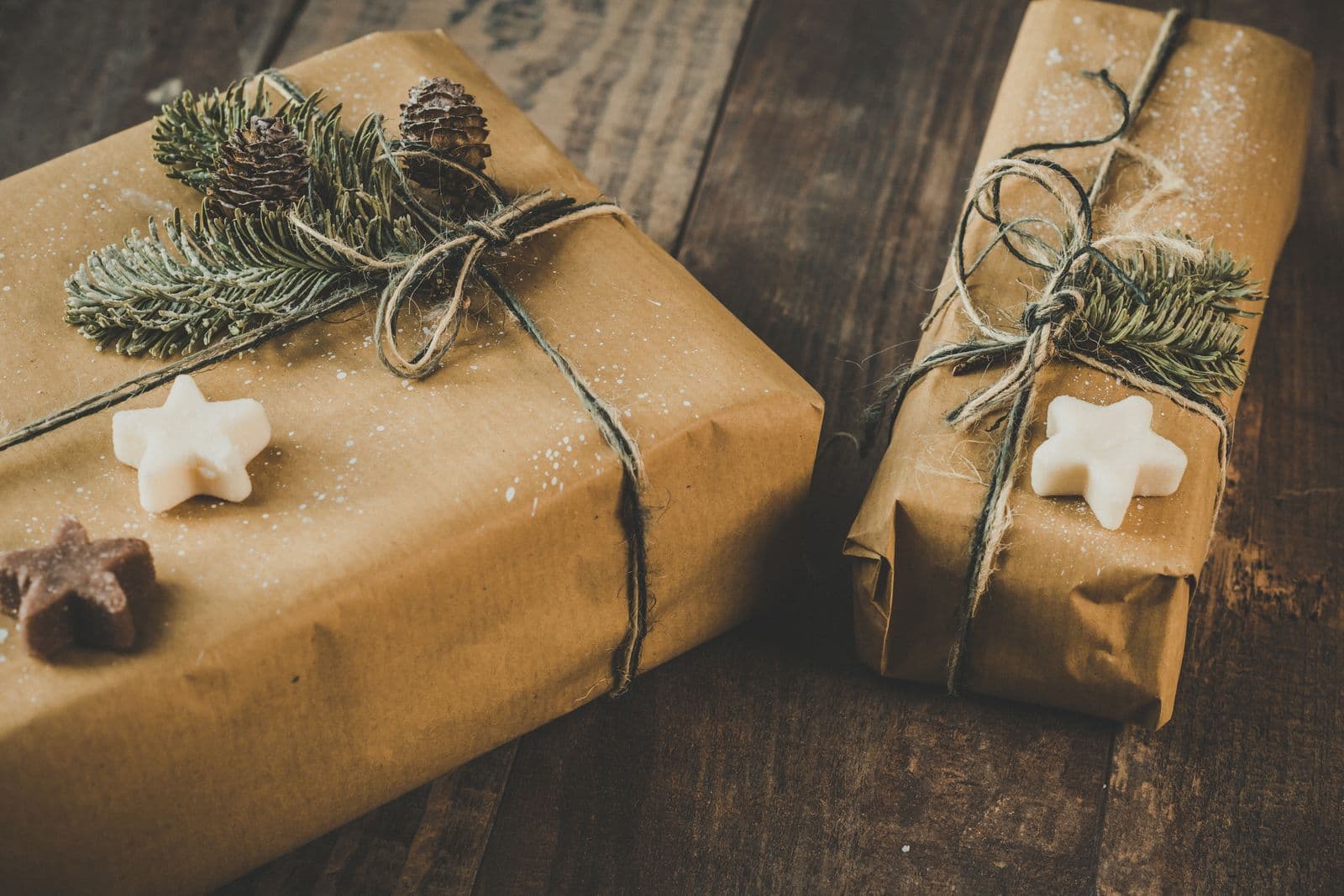 Two gifts wrapped in kraft paper and tied with jute twine, each finished with a cedar sprig and a small white star, resting on dark weathered wood