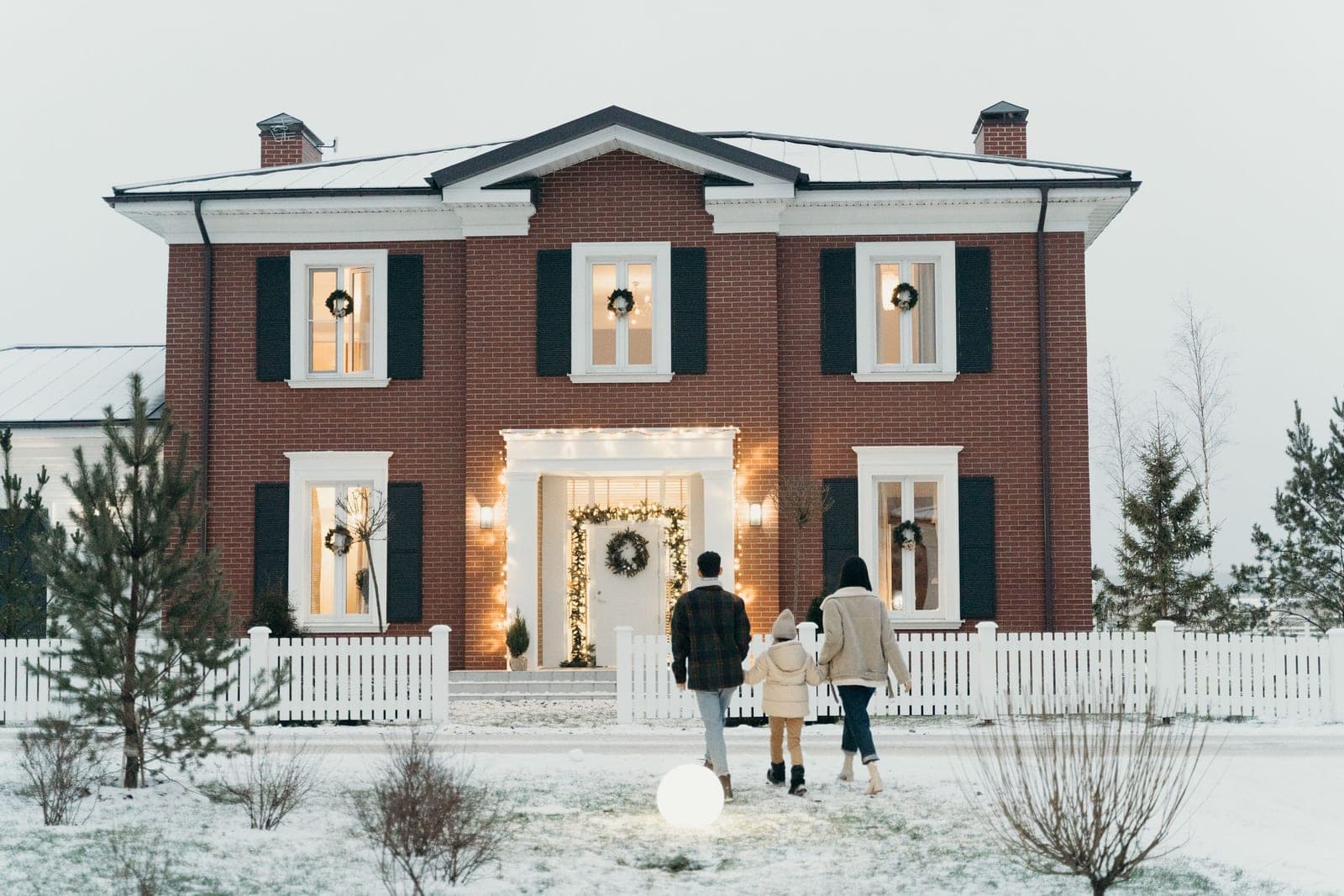 A family walking up a snowy front lawn toward a red brick colonial with a lit evergreen wreath in every window and warm string lights framing the white portico