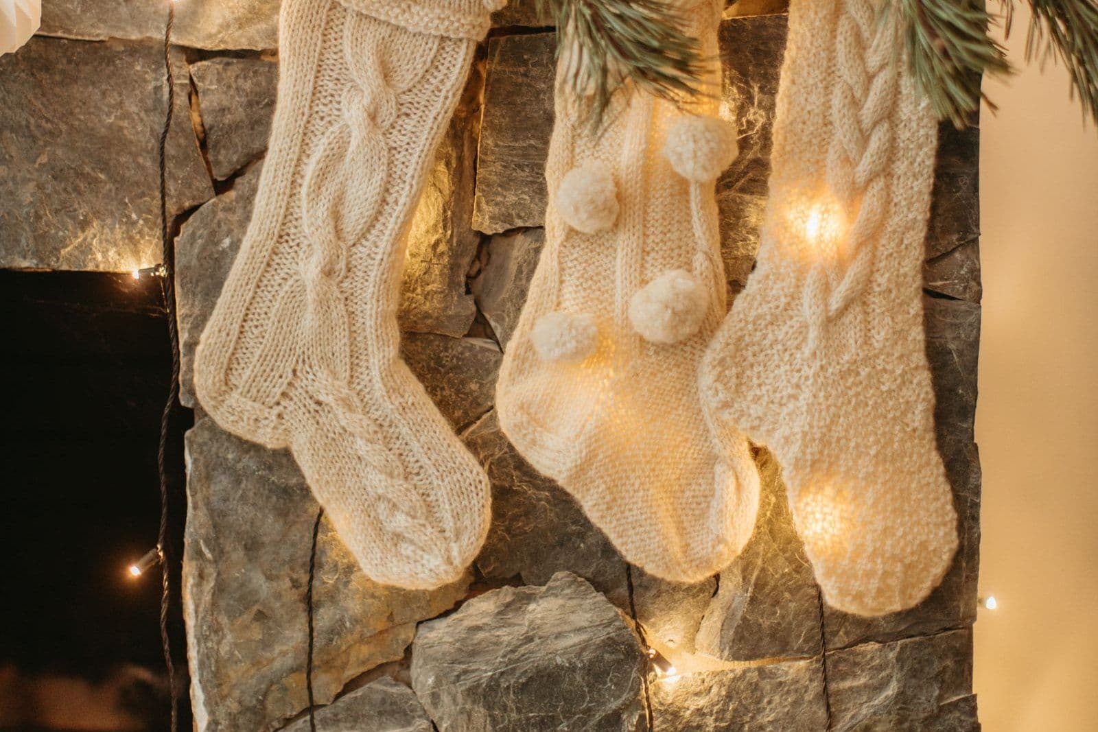 Cream cable-knit Christmas stockings hanging against a stone wall with warm fairy lights woven behind them