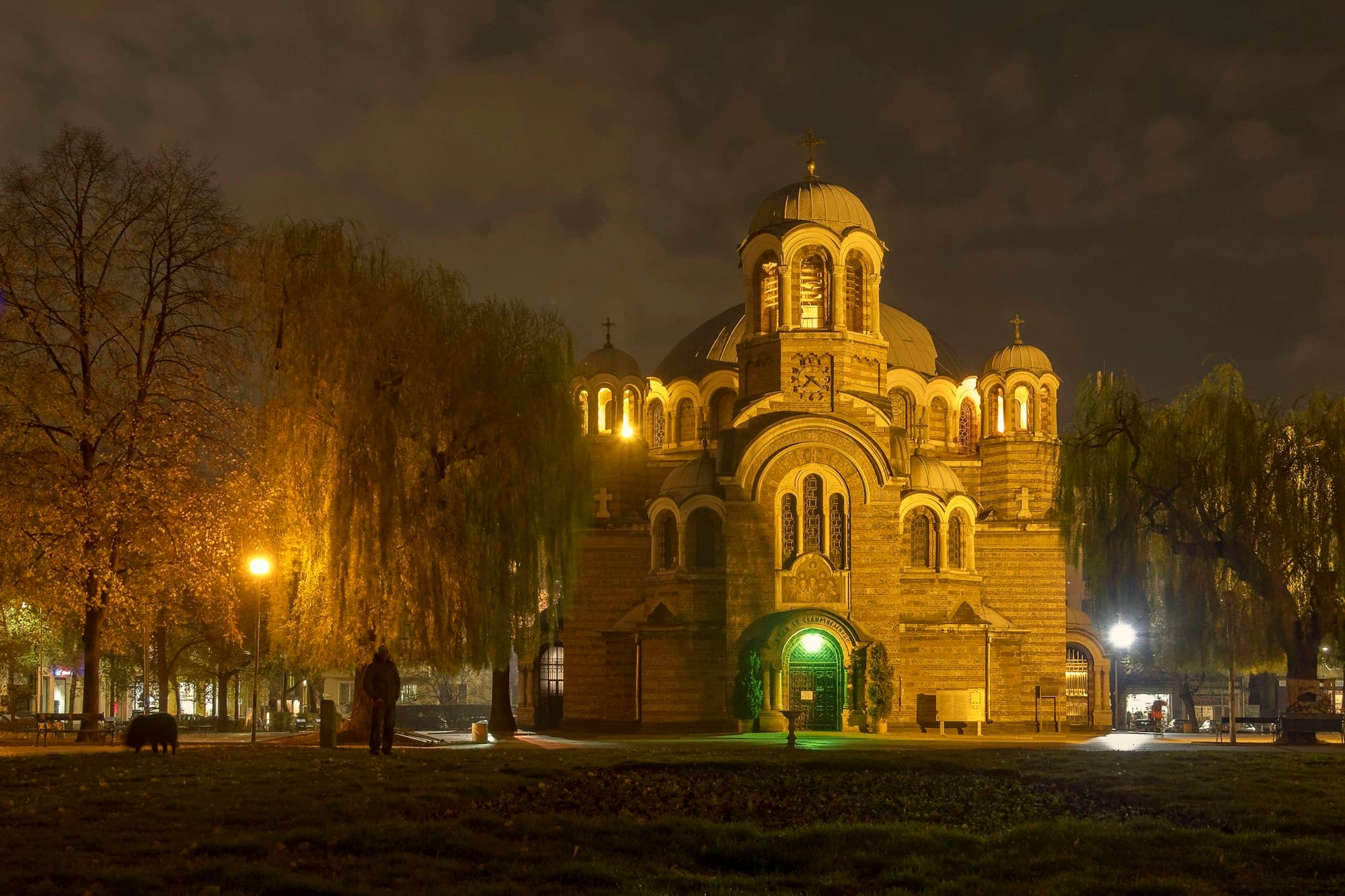 The Sveti Sedmochislenitsi Church in Sofia, Bulgaria glowing warm gold under a dark night sky, its Bulgarian Orthodox dome and arches lit from below, framed by bare willow trees over a quiet park
