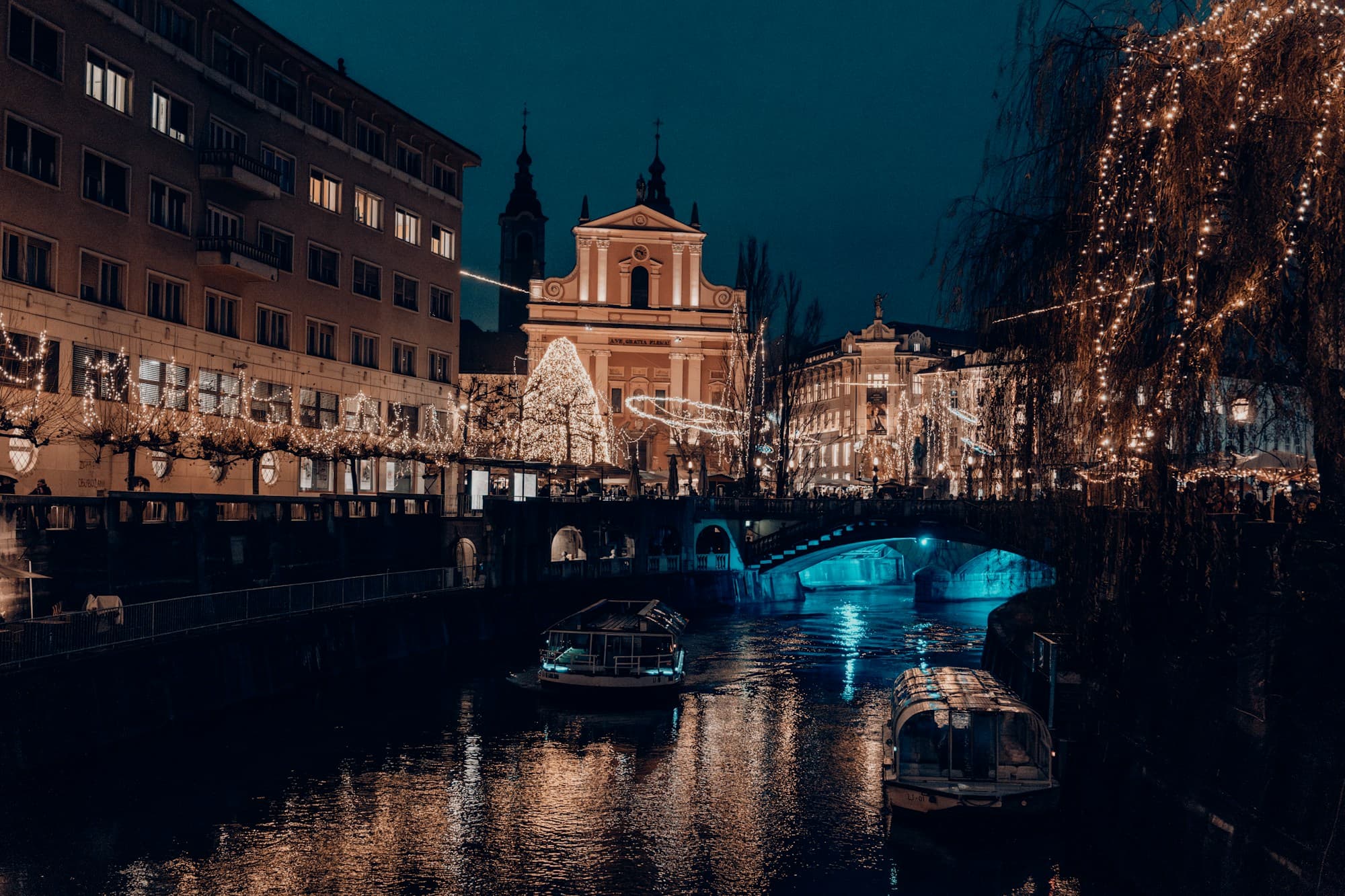 The Ljubljanica River in Ljubljana, Slovenia at night during Christmas, with the pink Franciscan Church of the Annunciation lit on Prešeren Square, festive lights strung along the riverbanks and cascading from a willow tree, and small boats on the water beneath an illuminated bridge