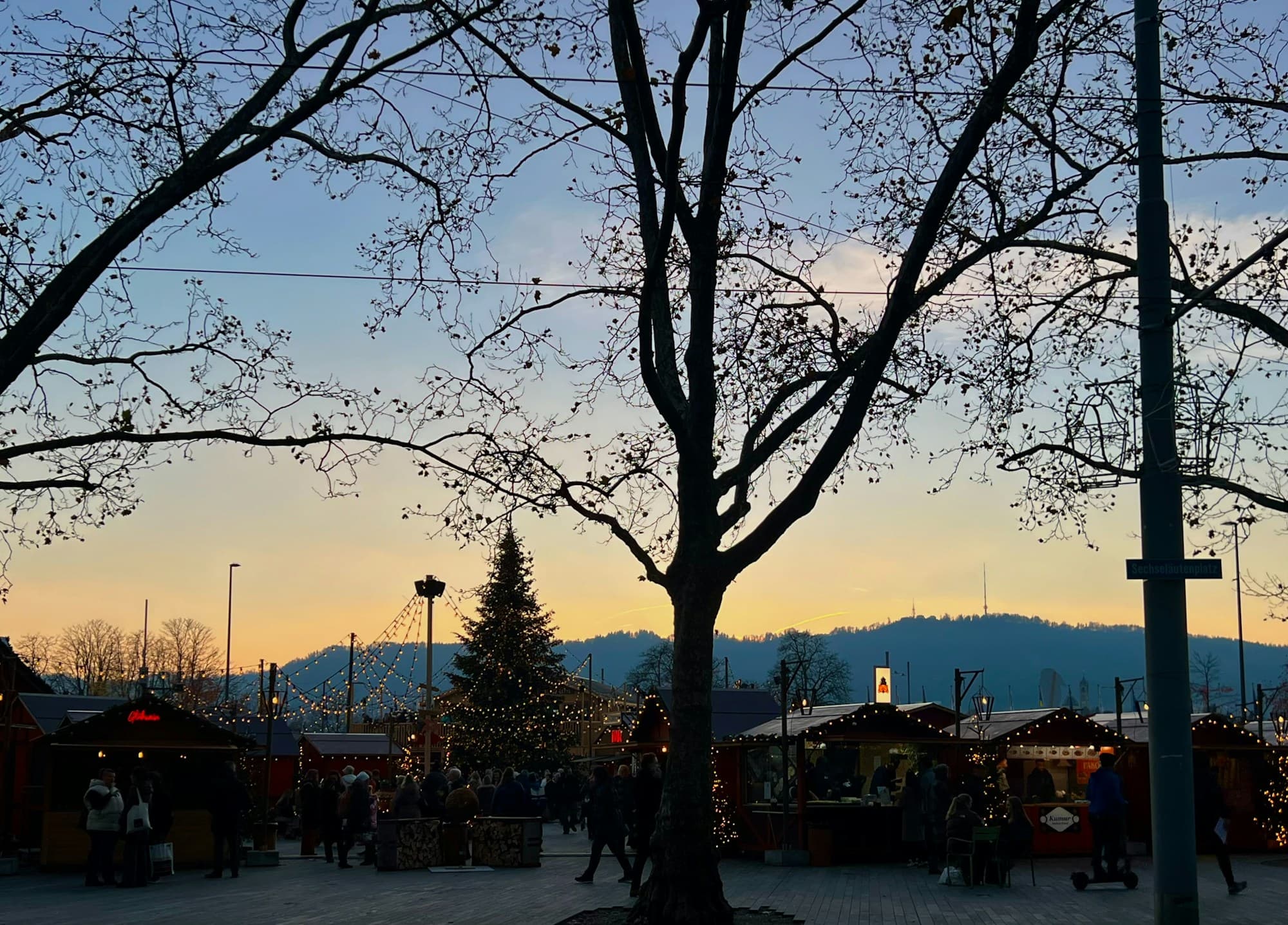 A Swiss Christmas market at golden hour, with a central illuminated Christmas tree, rows of wooden market stalls strung with warm white lights, the silhouette of a bare winter tree in the foreground, and alpine hills rising behind under a soft sunset sky